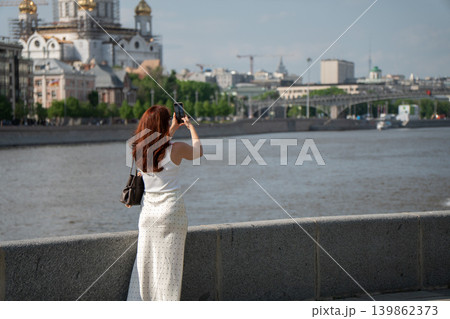 Red-haired woman photographs Cathedral of Christ the Saviour from Moscow River embankment under blue sky with city skyline, water view. Tourism, mobile photography, travel, city landmark, social media Red-haired woman photographs Cathedral of Christ the Saviour from Moscow River embankment under blue sky with city skyline, water view. Tourism, mobile photography, travel, city landmark, social media 139862373