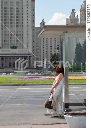 Red-haired woman leans against bus stop shelter with Moscow State University main building and wide city avenue. Tourism, lifestyle, city travel, urban architecture, social media Red-haired woman leans against bus stop shelter with Moscow State University main building and wide city avenue. Tourism, lifestyle, city travel, urban architecture, social media 139862379