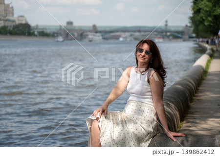 Red-haired woman in sunglasses sits on Gorky Park embankment beside Moscow River with bridge and city waterfront. Lifestyle, wellness branding, city leisure, travel advertising, summer recreation Red-haired woman in sunglasses sits on Gorky Park embankment beside Moscow River with bridge and city waterfront. Lifestyle, wellness branding, city leisure, travel advertising, summer recreation 139862412