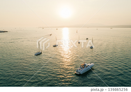 Aerial view of numerous sailing boats and a large motor yacht spread across the water during a vibrant golden sunset Aerial view of numerous sailing boats and a large motor yacht spread across the water during a vibrant golden sunset 139865256