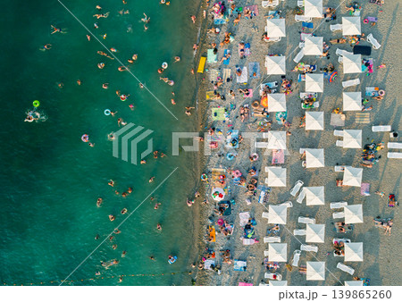 Numerous people enjoy a summer day at the beach. Swimmers cool off in the turquoise sea while others sunbathe under rows of white umbrellas and lounge chairs Numerous people enjoy a summer day at the beach. Swimmers cool off in the turquoise sea while others sunbathe under rows of white umbrellas and lounge chairs 139865260