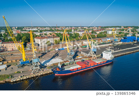 Aerial scene of an active industrial port. Yellow cranes load bulk materials into cargo ships docked at the waterfront Aerial scene of an active industrial port. Yellow cranes load bulk materials into cargo ships docked at the waterfront 139865346