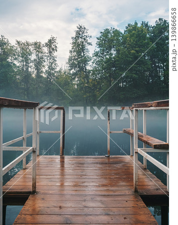 Wooden pier with metal railings extending into a misty lake in the early morning near a forest on a cloudy day 139866658