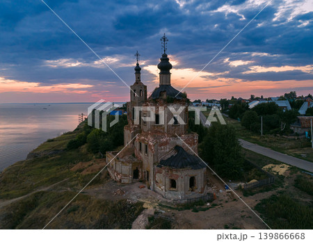 An old, crumbling Russian Orthodox church with two domes stands on a grassy cliff above a wide river during sunset An old, crumbling Russian Orthodox church with two domes stands on a grassy cliff above a wide river during sunset 139866668