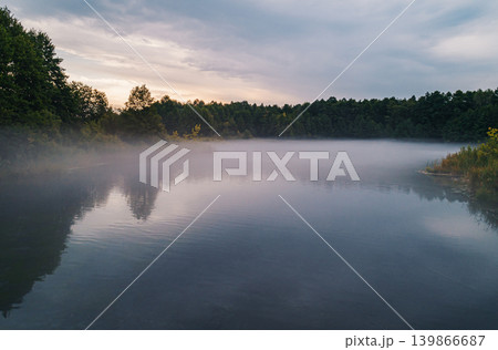 Serene morning mist covers peaceful lake surrounded by green forest. Serene morning mist covers peaceful lake surrounded by green forest. 139866687