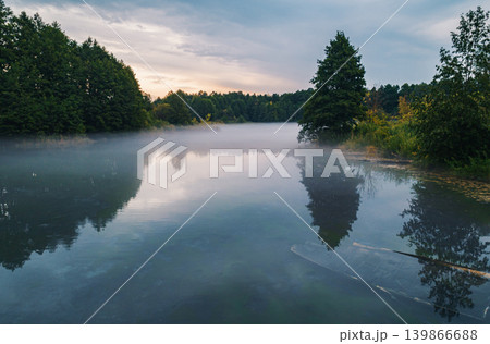 Serene morning mist covers peaceful lake surrounded by green forest. Serene morning mist covers peaceful lake surrounded by green forest. 139866688