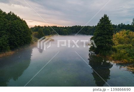Serene morning mist covers peaceful lake surrounded by green forest. Serene morning mist covers peaceful lake surrounded by green forest. 139866690
