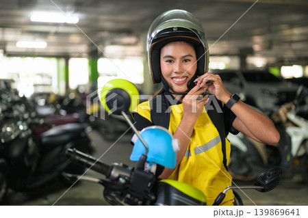 Smiling Female Delivery Courier Fastening Helmet in Parking Garage Smiling Female Delivery Courier Fastening Helmet in Parking Garage 139869641