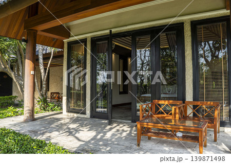 Tropical resort villa entrance in Vietnam with wooden bench and table on a shaded patio. Large glass doors, wooden ceiling, stone columns, and lush garden surroundings. Tropical resort villa entrance in Vietnam with wooden bench and table on a shaded patio. Large glass doors, wooden ceiling, stone columns, and lush garden surroundings. 139871498