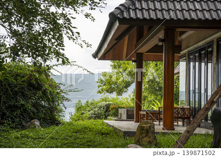 Luxury tropical villa in Vietnam with traditional tiled roof, wooden columns, and glass doors. Lush green garden, stone boulders, and stunning sea and mountain view. Luxury tropical villa in Vietnam with traditional tiled roof, wooden columns, and glass doors. Lush green garden, stone boulders, and stunning sea and mountain view. 139871502