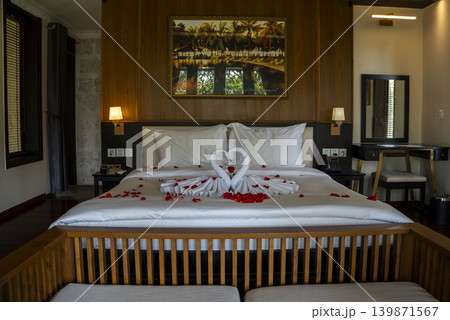 Romantic luxury hotel room in Vietnam with towel swans forming a heart shape and red rose petals on a king bed. Wooden decor, warm lighting, and tropical painting on the wall. Romantic luxury hotel room in Vietnam with towel swans forming a heart shape and red rose petals on a king bed. Wooden decor, warm lighting, and tropical painting on the wall. 139871567