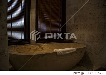 Stunning freestanding onyx stone bathtub with golden veining and white towel in a luxury resort bathroom in Vietnam. Dark wood blinds, marble walls, and warm ambient lighting. 139871572