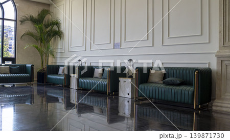 Luxurious lobby lounge with a row of green leather channel-tufted sofas, gold accents, marble side tables, and globe lamps. White wainscoting walls, reflective marble floor, and tropical palm tree. 139871730