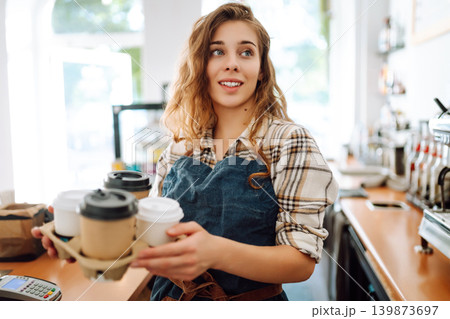 The young barista owner behind the bar gives out to-go orders. Business concept. Drinks to go. The young barista owner behind the bar gives out to-go orders. Business concept. Drinks to go. 139873697
