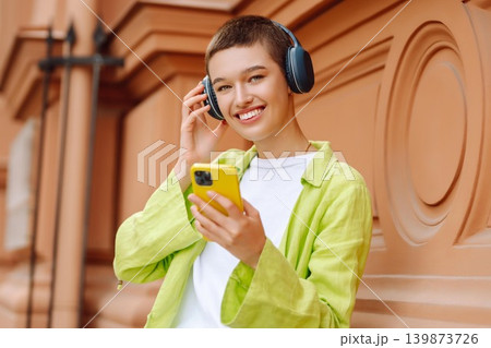 Woman wearing headphones posing outdoors, listens to music and walks along streets of a sunny city. Woman wearing headphones posing outdoors, listens to music and walks along streets of a sunny city. 139873726