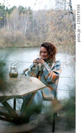 A sweet woman with curly hair drinks tea and relaxes on the terrace of her house on an autumn day. Health care, sincerity, a sense of balance and calm. 139874398