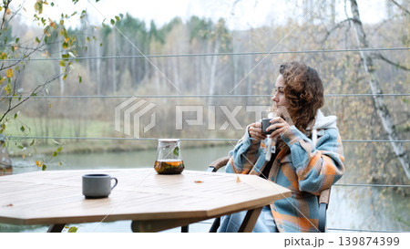 A sweet woman with curly hair drinks tea and relaxes on the terrace of her house on an autumn day. Health care, sincerity, a sense of balance and calm. 139874399