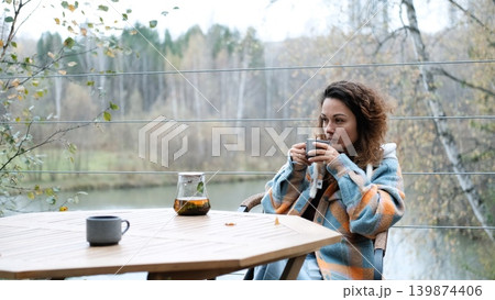 A sweet woman with curly hair drinks tea and relaxes on the terrace of her house on an autumn day. Health care, sincerity, a sense of balance and calm. 139874406