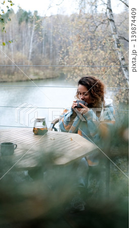 A sweet woman with curly hair drinks tea and relaxes on the terrace of her house on an autumn day. Health care, sincerity, a sense of balance and calm. A sweet woman with curly hair drinks tea and relaxes on the terrace of her house on an autumn day. Health care, sincerity, a sense of balance and calm. 139874409