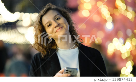 A beautiful girl holding a phone during a night walk through the city. Bokeh and lens flare effects on a blurred background. A beautiful girl holding a phone during a night walk through the city. Bokeh and lens flare effects on a blurred background. 139874415