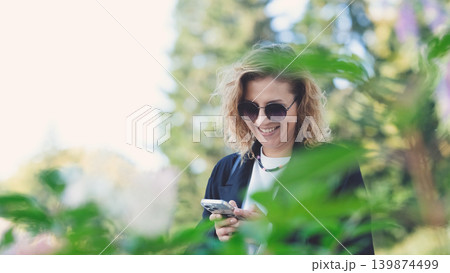 Caucasian woman with curly hair using her mobile phone outdoors. Close-up. Communication technology and modern lifestyle concept Caucasian woman with curly hair using her mobile phone outdoors. Close-up. Communication technology and modern lifestyle concept 139874499