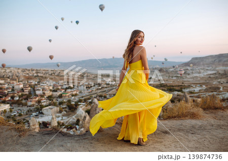 Woman stands on mountain with flying air balloons on background. Tourist Turkish region Cappadocia. Woman stands on mountain with flying air balloons on background. Tourist Turkish region Cappadocia. 139874736