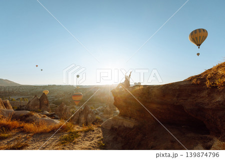 Happy girl in hat stands on mountain with flying air balloons on background. View from the back. Happy girl in hat stands on mountain with flying air balloons on background. View from the back. 139874796