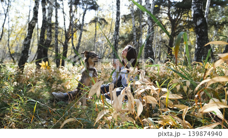 A cute young woman drinks tea from a thermos while walking her dog in the autumn forest. The woman enjoys the beauty of nature and the connection with her pets 139874940