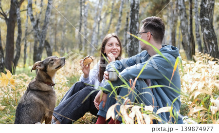 A married couple relaxes in nature during a walk with their beloved dog in an autumn forest. The loving woman and man sit in the tall yellow grass, in the fern leaves, with the dog next to them. A married couple relaxes in nature during a walk with their beloved dog in an autumn forest. The loving woman and man sit in the tall yellow grass, in the fern leaves, with the dog next to them. 139874977