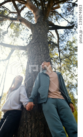 Lovers boy and girl standing near a big pine tree during a walk on a sunny autumn day Lovers boy and girl standing near a big pine tree during a walk on a sunny autumn day 139874983