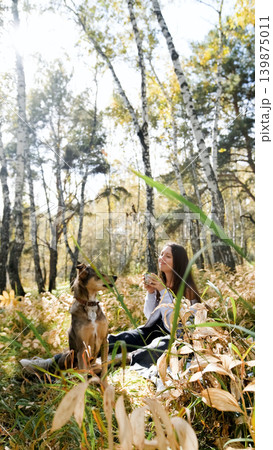 A sweet young woman with her dog enjoys the beauty of nature and the bond with her pets. The woman drinks tea from a thermos and eats buns A sweet young woman with her dog enjoys the beauty of nature and the bond with her pets. The woman drinks tea from a thermos and eats buns 139875011