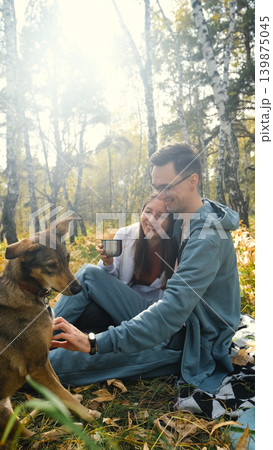 A married couple relaxes in nature during a walk with their beloved dog in an autumn forest. The loving woman and man sit in the tall yellow grass, in the fern leaves, with the dog next to them. A married couple relaxes in nature during a walk with their beloved dog in an autumn forest. The loving woman and man sit in the tall yellow grass, in the fern leaves, with the dog next to them. 139875045