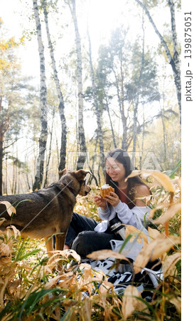 Cute young woman with her dog enjoying the beauty of nature and bonding with her pets. Woman eating bun and feeding her dog during outdoor picnic in autumn forest. Cute young woman with her dog enjoying the beauty of nature and bonding with her pets. Woman eating bun and feeding her dog during outdoor picnic in autumn forest. 139875051