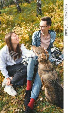 A married couple relaxes in nature during a walk with their beloved dog in an autumn forest. The loving woman and man sit in the tall yellow grass, in the fern leaves, with the dog next to them. A married couple relaxes in nature during a walk with their beloved dog in an autumn forest. The loving woman and man sit in the tall yellow grass, in the fern leaves, with the dog next to them. 139875585