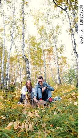 A married couple relaxes in nature during a walk with their beloved dog in an autumn forest. The loving woman and man sit in the tall yellow grass, in the fern leaves, with the dog next to them. A married couple relaxes in nature during a walk with their beloved dog in an autumn forest. The loving woman and man sit in the tall yellow grass, in the fern leaves, with the dog next to them. 139875609