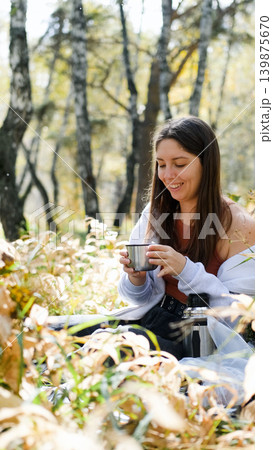 Cute young woman enjoying the beauty of nature, drinking tea from a thermos in the autumn forest Cute young woman enjoying the beauty of nature, drinking tea from a thermos in the autumn forest 139875670