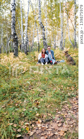 A married couple relaxes in nature during a walk with their beloved dog in an autumn forest. The loving woman and man sit in the tall yellow grass, in the fern leaves, with the dog next to them. A married couple relaxes in nature during a walk with their beloved dog in an autumn forest. The loving woman and man sit in the tall yellow grass, in the fern leaves, with the dog next to them. 139875979