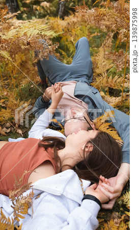A woman and a man in love lie in tall yellow grass, in fern leaves. The married couple relaxes in nature during a walk in the autumn forest. 139875998
