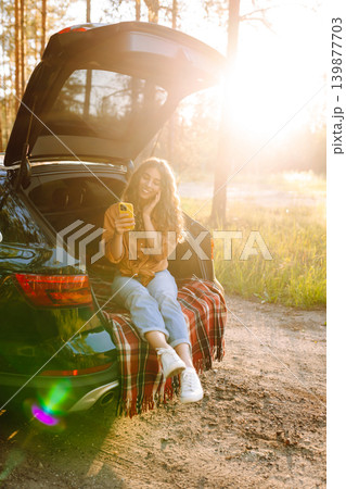 Young curly woman in stylish clothes in the trunk of a car in the forest and uses the phone. 139877703
