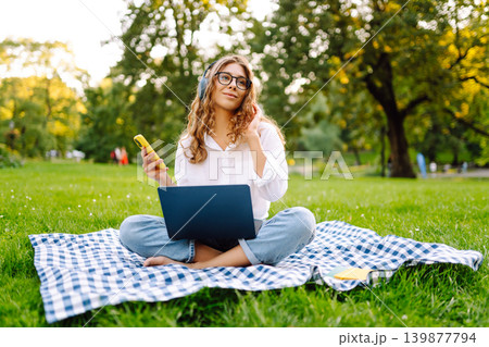 Woman with a laptop and headphones sitting on green grass in park studying, working. Music concept. 139877794