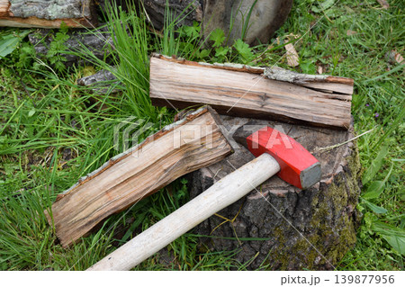 A red-handled axe rests on a wooden stump next to split logs. The scene is set in a grassy area with scattered wood pieces. A red-handled axe rests on a wooden stump next to split logs. The scene is set in a grassy area with scattered wood pieces. 139877956