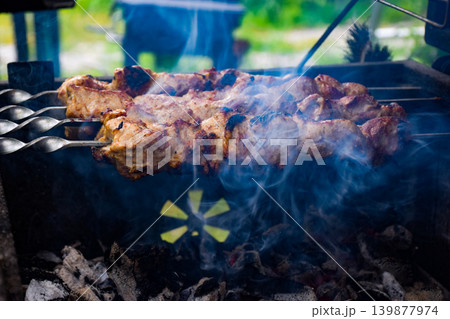 Grilled meat skewers cooking over an open flame. Smoke rises from the barbecue, creating a rustic outdoor cooking scene. Grilled meat skewers cooking over an open flame. Smoke rises from the barbecue, creating a rustic outdoor cooking scene. 139877974