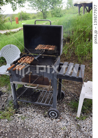 A charcoal grill with sausages cooking on it. The grill is placed outdoors on a gravel surface, surrounded by green grass and trees. A charcoal grill with sausages cooking on it. The grill is placed outdoors on a gravel surface, surrounded by green grass and trees. 139877977