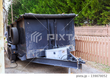 Trash trailer is parked next to fence in residential space with green trees nearby. Trash trailer is parked next to fence in residential space with green trees nearby. 139878052