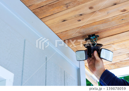 Handy man holds outdoor security lights while preparing to install them on wooden ceiling at their home. 139878059