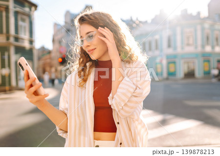 Optimistic young woman looking at camera with smile while taking selfie on city street. 139878213