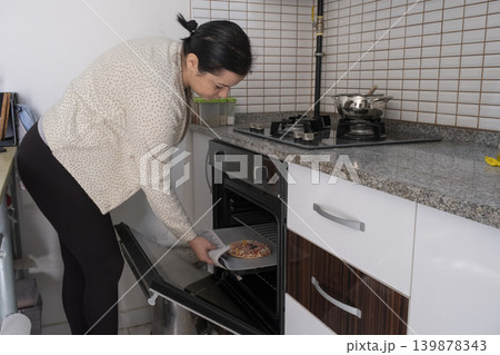 Woman Putting Frozen Pizza into Oven in Kitchen. Woman Putting Frozen Pizza into Oven in Kitchen. 139878343
