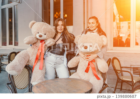 Women friendship bears holding giant plush toys at a sunny cafe terrace while smiling together during golden hour Women friendship bears holding giant plush toys at a sunny cafe terrace while smiling together during golden hour 139880687