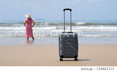 Suitcase on sandy beach in foreground while young woman in pink dress stands in calm sea water in background enjoying vacation. Holiday arrival concept Suitcase on sandy beach in foreground while young woman in pink dress stands in calm sea water in background enjoying vacation. Holiday arrival concept 139881513