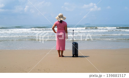 Rear view of young woman in pink dress and straw hat standing next to suitcase on beach gazing at the sea enjoying her vacation. Relaxation concept 139881517